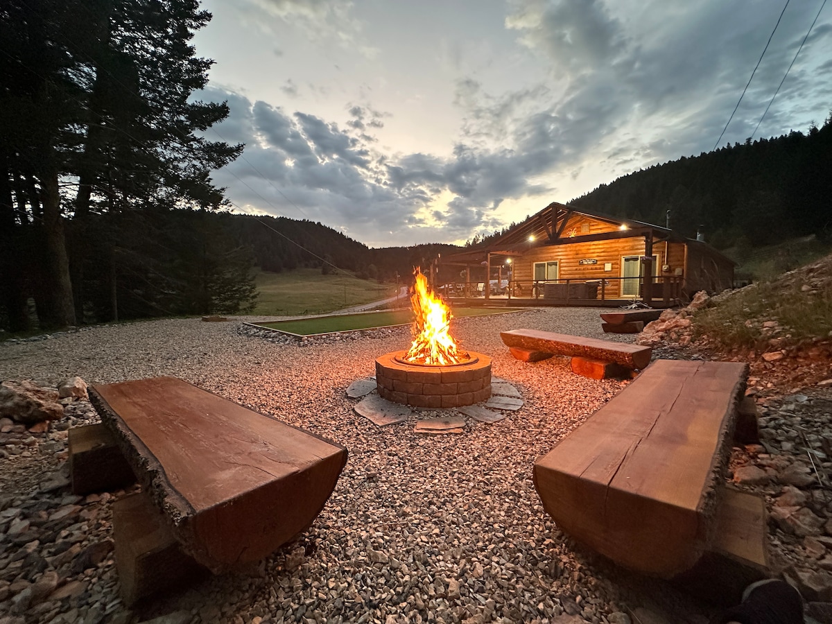 An outdoor fire pit is surrounded by two large wooden benches, set on gravel. The cabin is visible in the background, framed by trees and rolling hills. A sunset sky adds a warm glow to the scene, enhancing the peaceful ambiance.