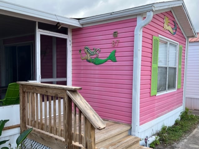 A charming pink cottage features colorful mermaid wall art. The front porch is accessible by a wooden ramp, leading to a door with green shutters and a large window. A small area of greenery is visible near the entrance.