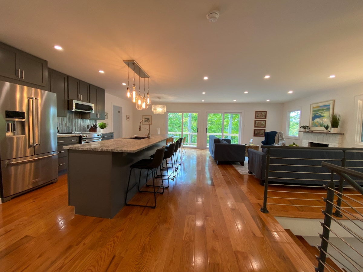 An open-concept kitchen and living area is showcased with modern appliances and a spacious island. The kitchen features dark cabinetry and stainless steel fixtures. Large glass doors reveal views of greenery outside, while ample natural light brightens the hardwood floors and comfortable furnishings.