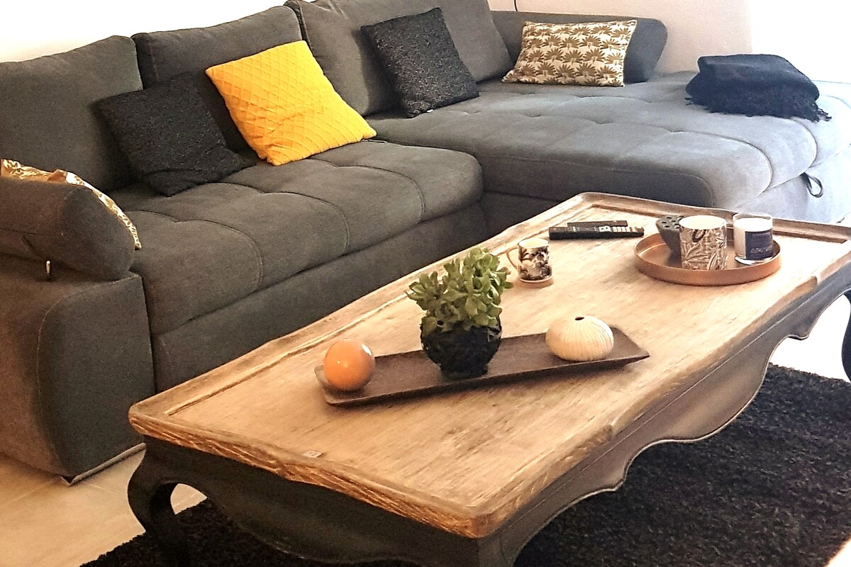 A spacious living area features a large, modern grey sectional sofa adorned with decorative pillows in various patterns. A rustic wooden coffee table is placed centrally, embellished with a small succulent plant, decorative items, and a remote control.