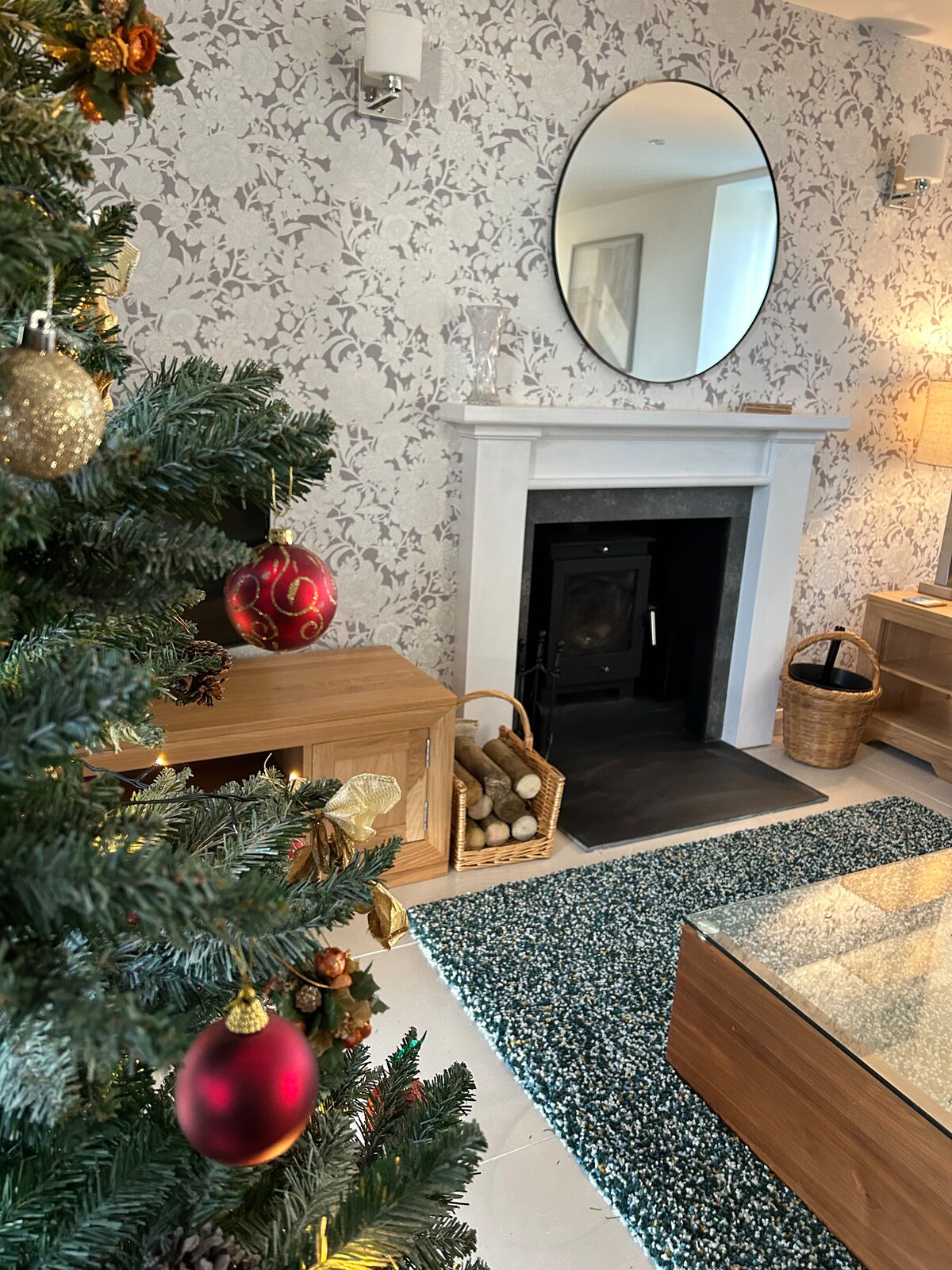 The living area features a decorative fireplace with a modern wood-burning stove, framed by a round mirror. A wooden side unit displays a basket of logs. A festive Christmas tree adds seasonal décor, while a textured area rug complements the light-colored furnishings.