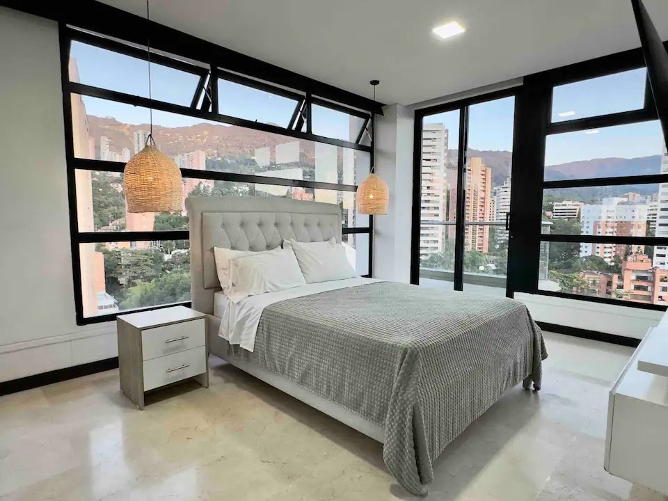 A spacious bedroom features a large, stylish bed with a tufted headboard. Natural light pours in through floor-to-ceiling windows, offering views of the city and mountains. Two woven pendant lights hang above the bed, and a bedside table complements the modern design.