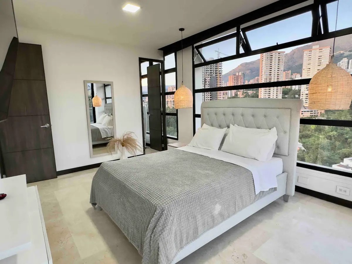 A spacious sleeping area features a comfortable bed with a light grey quilt. Large windows provide ample natural light along with a stunning view of the city and mountains. A decorative mirror stands adjacent to the bed, and pendant lights hang from the ceiling.