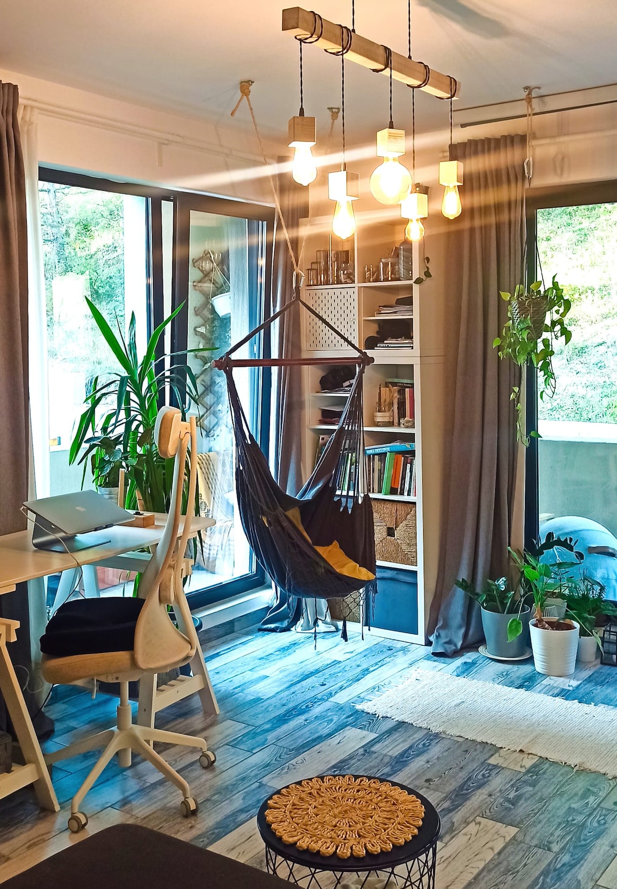 A cozy living space features a stylish hammock suspended from the ceiling, accompanied by a desk with a laptop. Floor-to-ceiling windows allow natural light to illuminate the room, where indoor plants and a bookshelf add to the inviting atmosphere. Warm light bulbs create a welcoming glow.