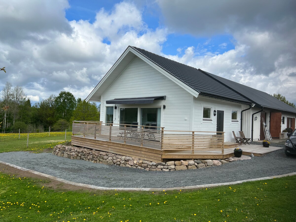 The exterior of the house is presented, featuring a modern design with a sloped roof and large windows. A wooden deck wraps around the front, accompanied by a stoned pathway. Lush greenery surrounds the property, and a gravel driveway leads to parked vehicles.