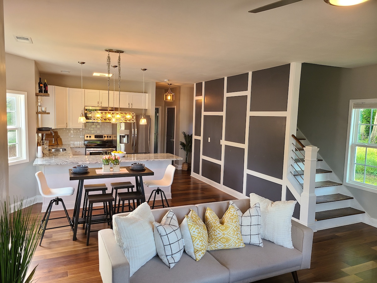 An open-concept living and dining area features a stylish dining table set for four, accompanied by modern black chairs. A well-equipped kitchen is visible in the background, showcasing white cabinetry and stainless steel appliances. Natural light fills the space through large windows.