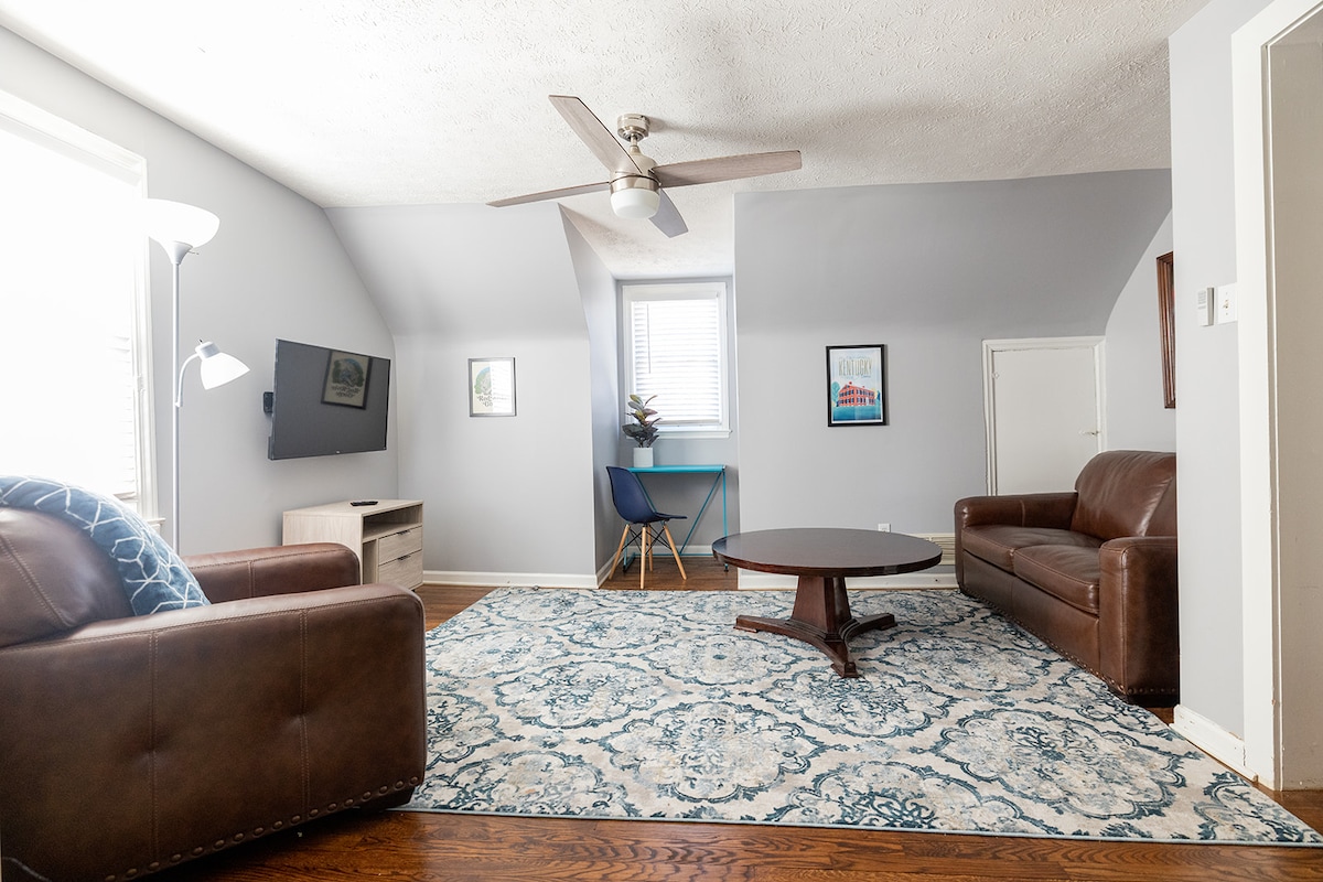 The living room is furnished with a large round coffee table at the center, two brown leather seating options, and a blue accent chair. A flat-screen television is mounted on the wall, and natural light enters through the windows, enhancing the space's openness.