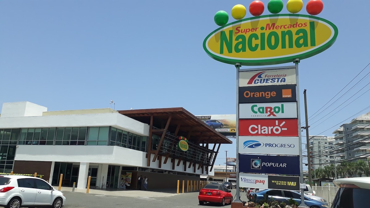The image displays the exterior of Super Mercados Nacional, featuring a modern architectural design with a wooden overhang. A colorful sign with large round lights at the top is visible, along with various brand logos on a vertical sign, indicating services available. The parking area includes several vehicles.