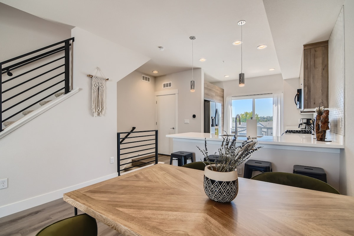 An open living area showcases a wooden dining table surrounded by comfortable green chairs. Bright light enters through large windows, highlighting a sleek kitchen in the background. A staircase with metal railings leads to the upper level, adding a modern touch to the space.