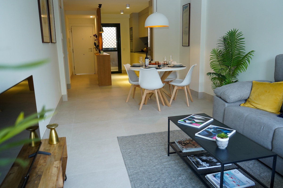 A bright and spacious living area is shown, featuring a comfortable sofa paired with decorative cushions. A wooden coffee table is centered, displaying magazines and a small plant. A dining table with white chairs is set for meals, with a kitchen area visible in the background.