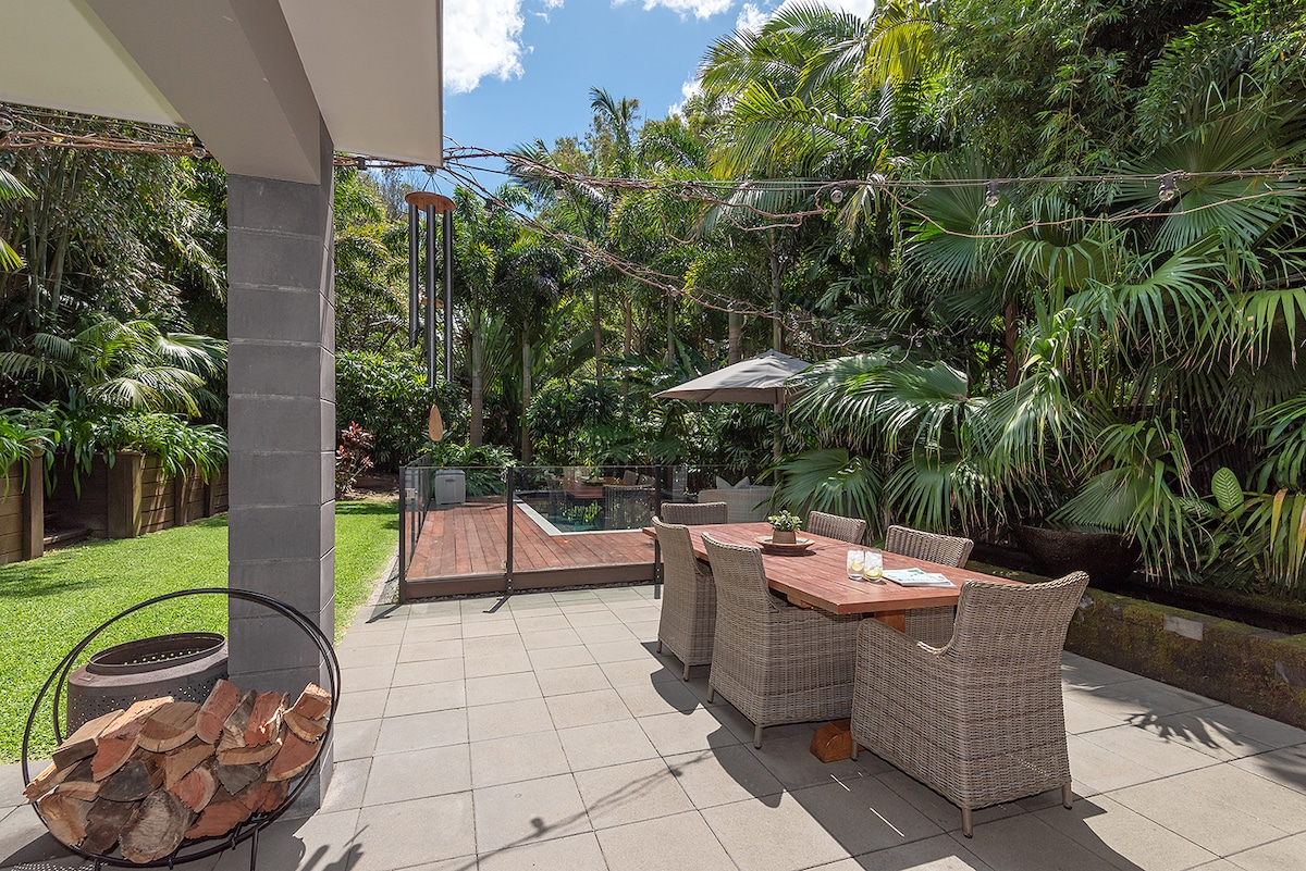 An outdoor dining area is surrounded by tropical greenery, featuring a wooden table and wicker chairs. A shaded umbrella provides comfort, while a spacious deck with a heated pool is visible in the background, inviting relaxing moments in a tranquil setting.