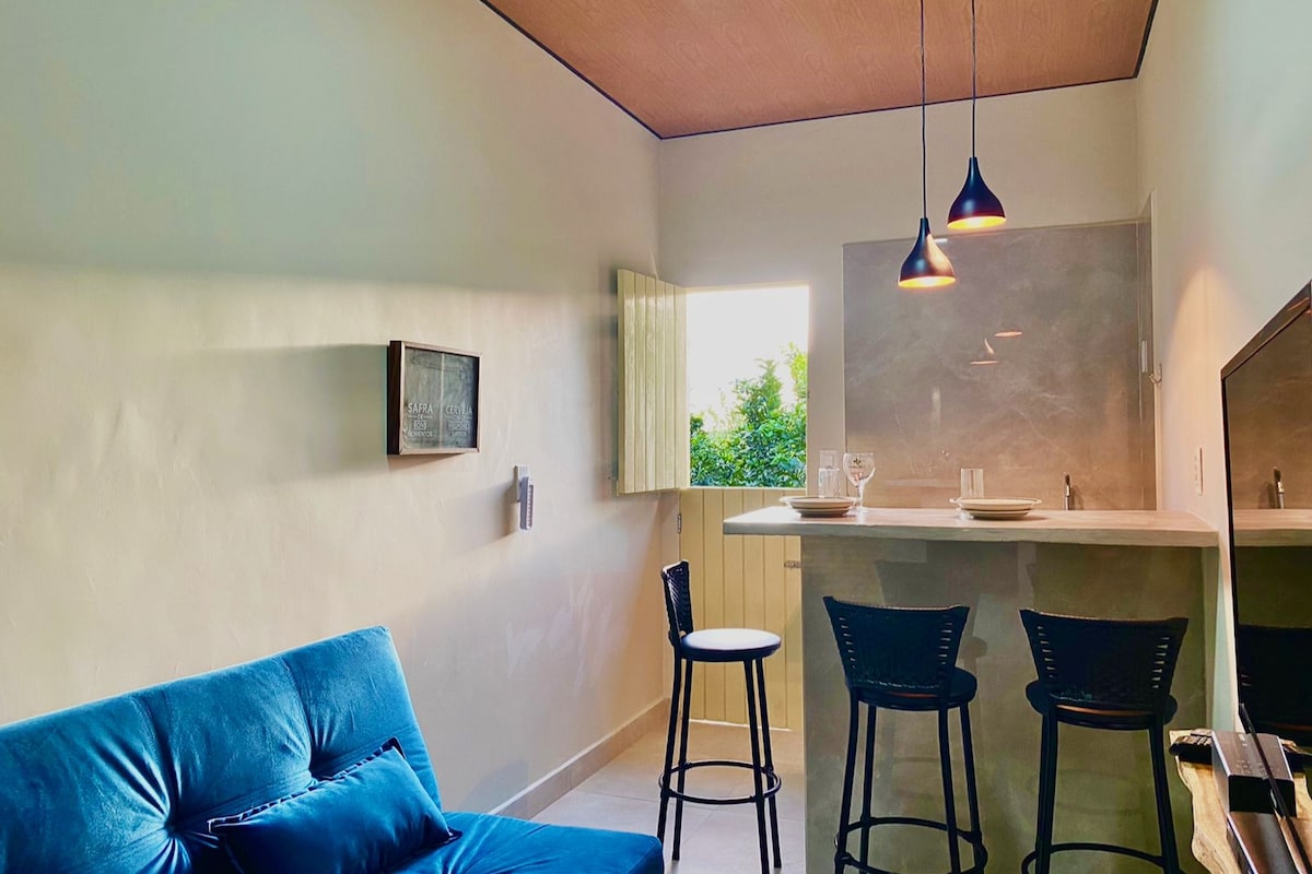A comfortable seating area features a blue sofa alongside a high table with black bar stools. Natural light enters through a window, illuminating the modern kitchen setup in the background, which includes simple countertop amenities.