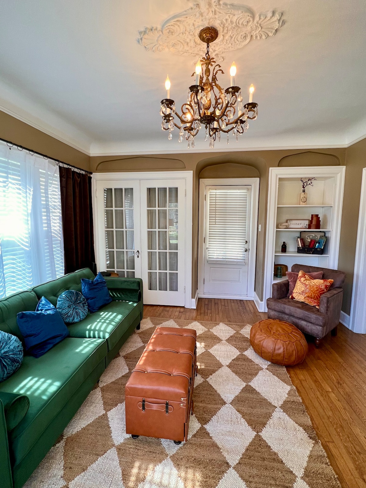 A stylish living area features a green sofa adorned with colorful cushions. A wooden ottoman is positioned in front, complemented by a round pouf. A chandelier hangs from the ceiling, enhancing the vintage charm. Light floods through large, sheer-draped windows, illuminating the space.
