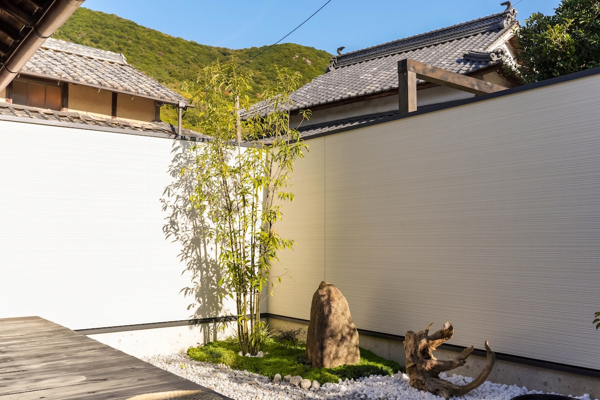 A tranquil garden space features a bamboo plant and a smooth stone surrounded by white pebbles. The clean, minimalist design is complemented by surrounding walls that enhance the sense of peace. Natural sunlight casts gentle shadows, highlighting the serene atmosphere.