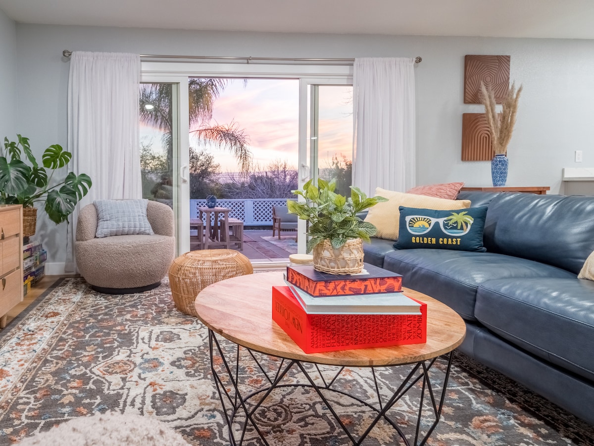 A modern living area features a comfortable blue leather sofa and a light, textured armchair. A wooden coffee table with decorative items is centered on a patterned rug. Large sliding glass doors provide a view of the sunset and access to the outdoor space.