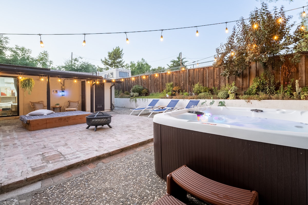 An outdoor entertainment area is showcased, featuring a hot tub surrounded by lounge seating and illuminated by string lights. A brick patio area includes a fire pit, while sun loungers are positioned along the edge of a garden filled with greenery.