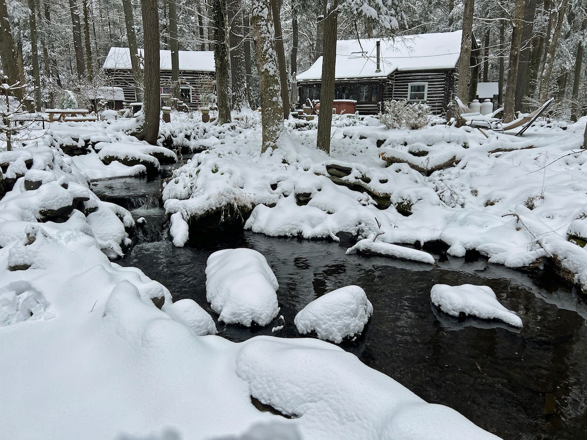 A serene winter scene showcases a flowing creek, partially covered in a blanket of snow. Snow-dusted rocks and banks line the waterway, with a view of rustic log cabins nestled among snow-laden trees in the background.