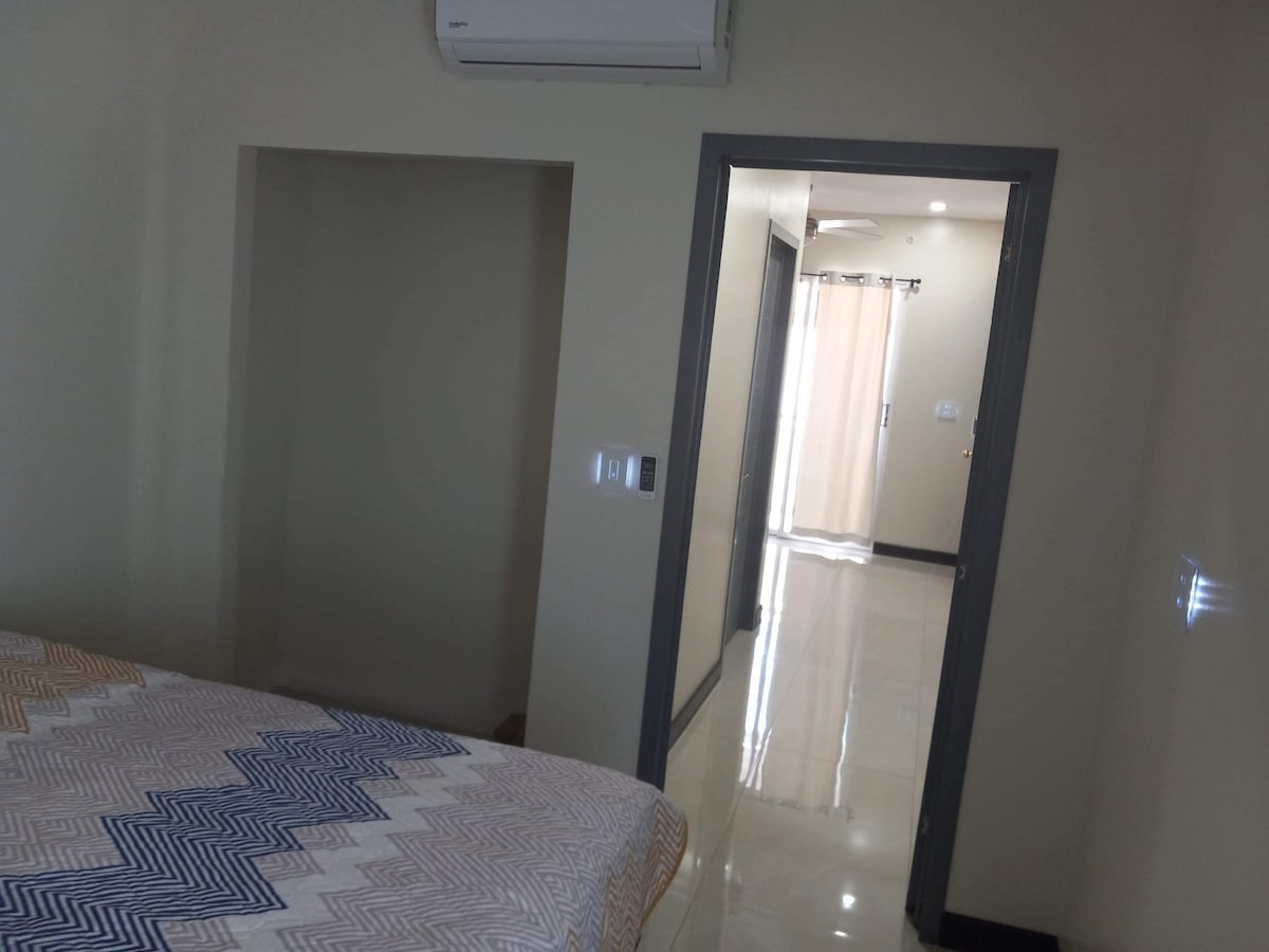 A spacious bedroom is depicted with a patterned bedspread. A doorway leads to a well-lit hallway featuring smooth tiled flooring and natural light flowing through sheer curtains. Air conditioning is installed on the wall for comfort.
