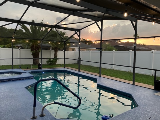A private pool and spa area is enclosed by a screen, offering views of a serene sunset through palm trees. The pool features a textured surface and a safety railing, with vibrant water reflecting the evening sky. A lush green lawn surrounds the space, contributing to the tranquil environment.