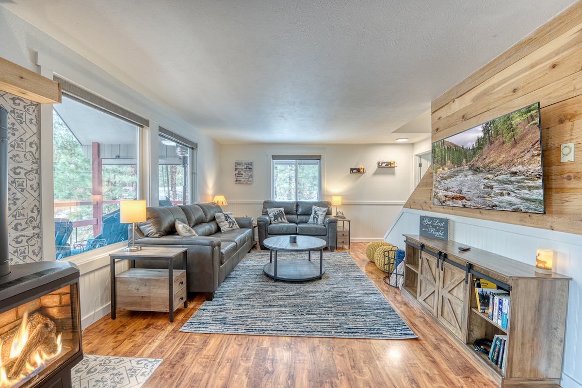 A comfortable living area is showcased with a plush seating arrangement and a circular coffee table. A fireplace adds warmth, while natural light filters through large windows. An entertainment center holds a television, and a subtle area rug complements the wooden flooring.