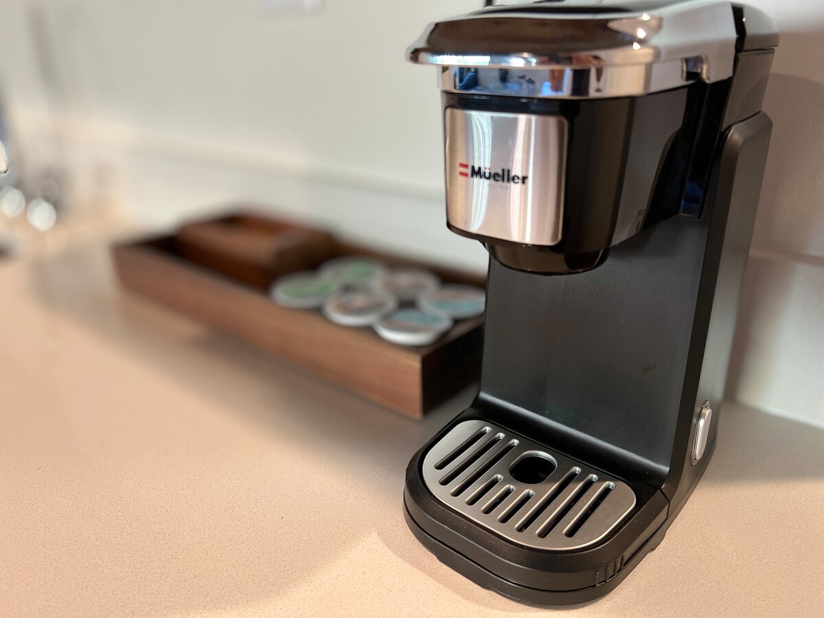 A single-serve coffee maker is set on a light-colored countertop. A wooden tray with various coffee pods is positioned nearby, offering a selection of flavors. The appliance features a sleek design and a metallic finish, emphasizing modern convenience.