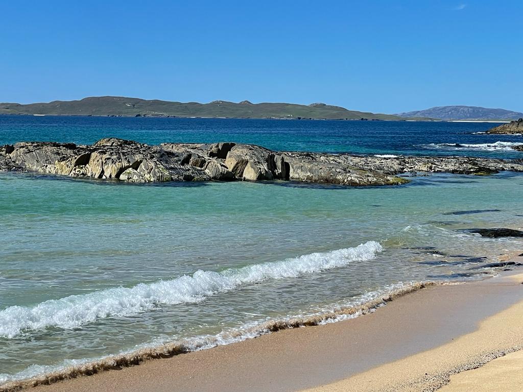 The image captures a serene beach scene featuring gentle waves lapping against smooth sandy shores. Rocky formations are visible in the water, with a backdrop of distant hills and clear blue skies, creating a calm coastal landscape.