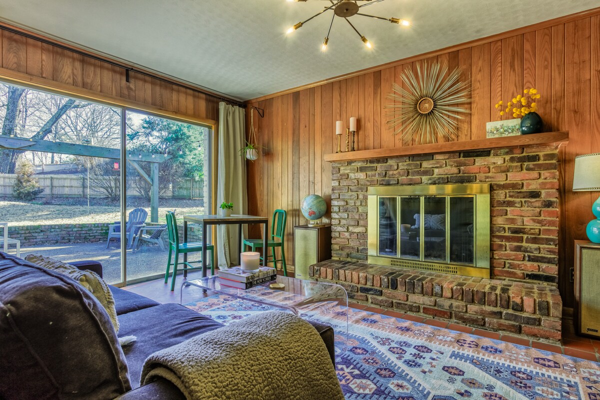 A warm living room is showcased with wooden paneling and a brick fireplace at its center. A large window allows natural light to fill the space, revealing a view of the outdoor area. A patterned rug lies under a coffee table, complemented by green chairs and decorative elements.