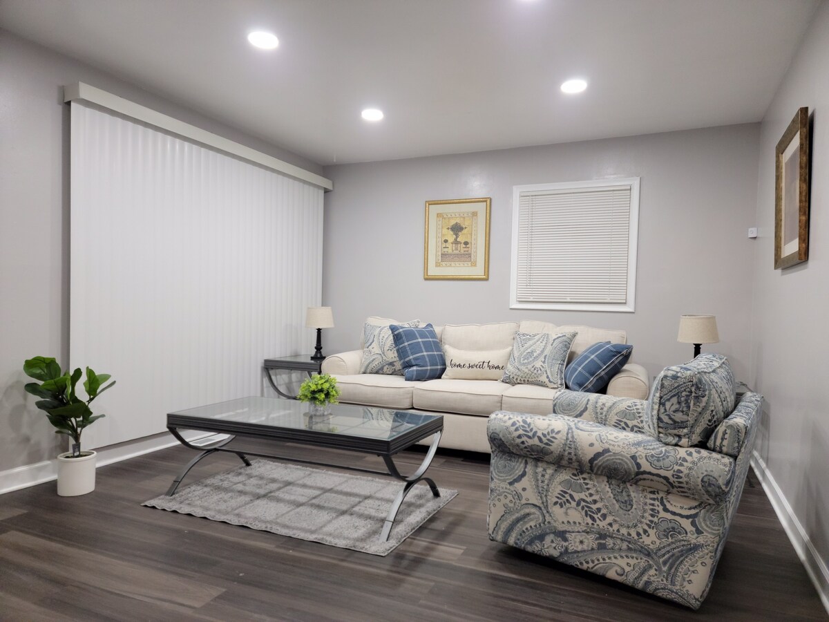 A living room is presented with a comfortable couch adorned with blue and white cushions. A patterned armchair complements the space, while a glass coffee table is centrally placed. Soft lighting enhances the room's ambiance, and window treatments provide light control.