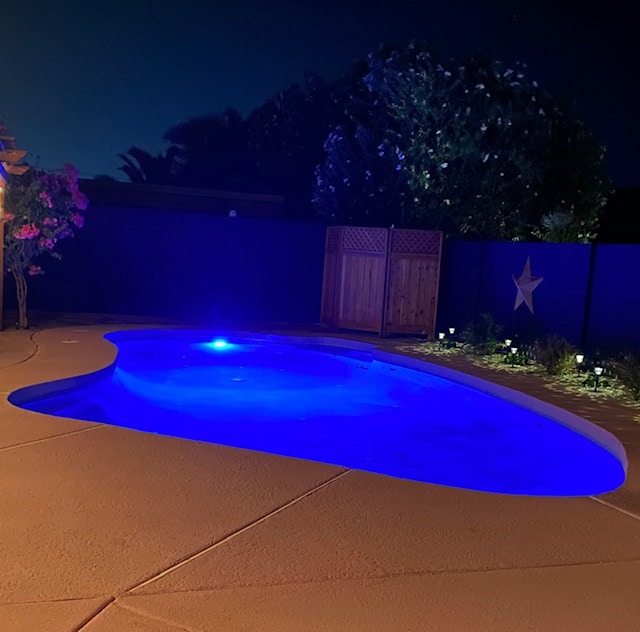 A serene pool area is illuminated with soft blue lights, creating a calming ambiance at night. Surrounding the pool are landscaped features and walls, providing a sense of privacy. The pool's organic shape complements the tranquil setting, with shadows of nearby trees softly visible.