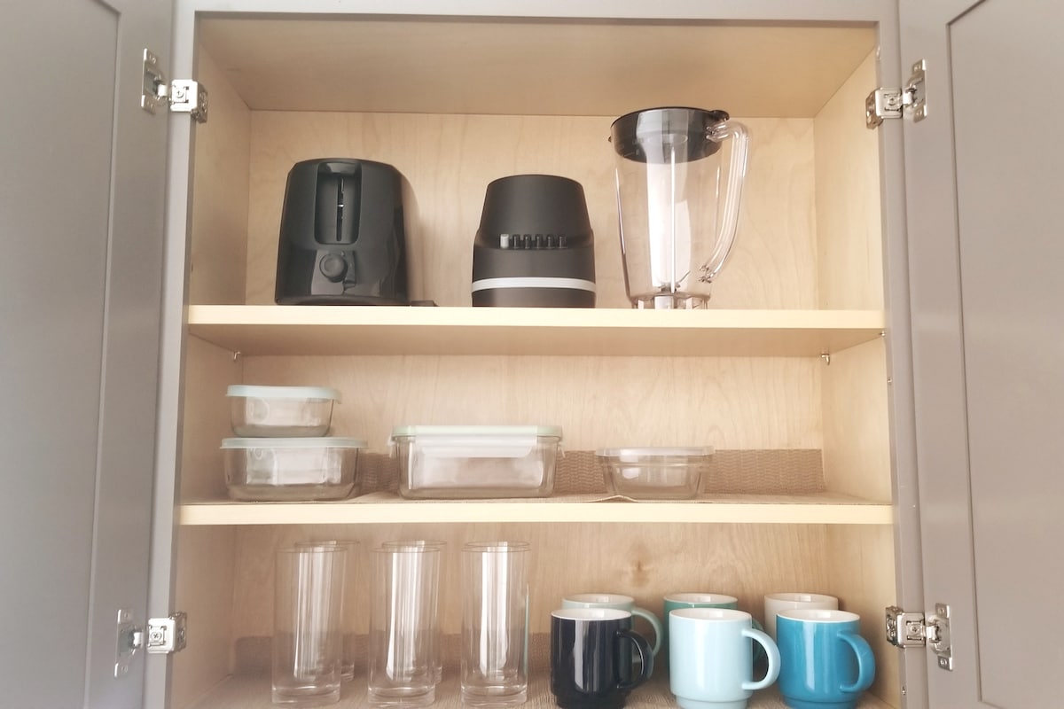 A kitchen cabinet is organized with kitchen appliances and glassware. The top shelf holds a toaster, coffee maker, and blender. The second shelf features various glass storage containers. The bottom shelf contains clear drinking glasses and a collection of colorful mugs.