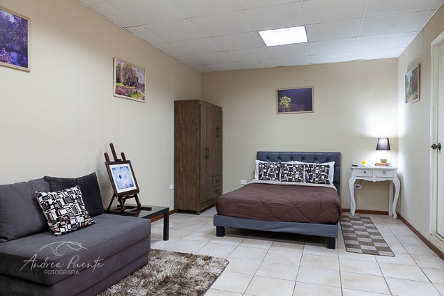 A spacious studio room features a double bed with a brown quilt, accompanied by decorative pillows. A comfortable couch is positioned beside a small side table. Art prints adorn the walls, while tile flooring offers a clean and practical aesthetic.