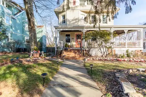 The exterior of a renovated Victorian home is shown, featuring a charming porch and landscaped yard. A paved walkway leads to the front steps, bordered by garden beds with seasonal plants. Trees provide shade, enhancing the inviting atmosphere of the property.