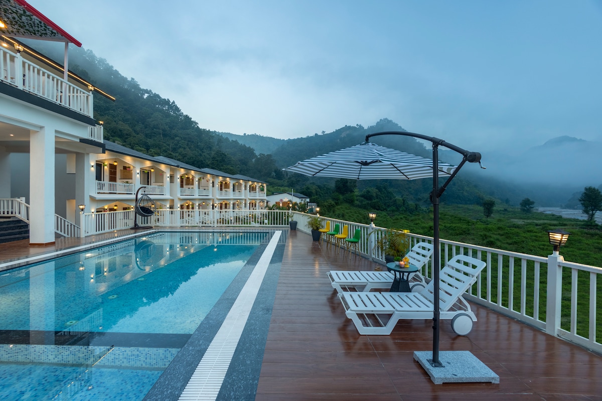 The image captures a serene swimming pool bordered by a wooden deck. Several lounge chairs are positioned for relaxation under a large umbrella, while misty mountains rise in the background. The tranquil atmosphere is complemented by the greenery surrounding the property.