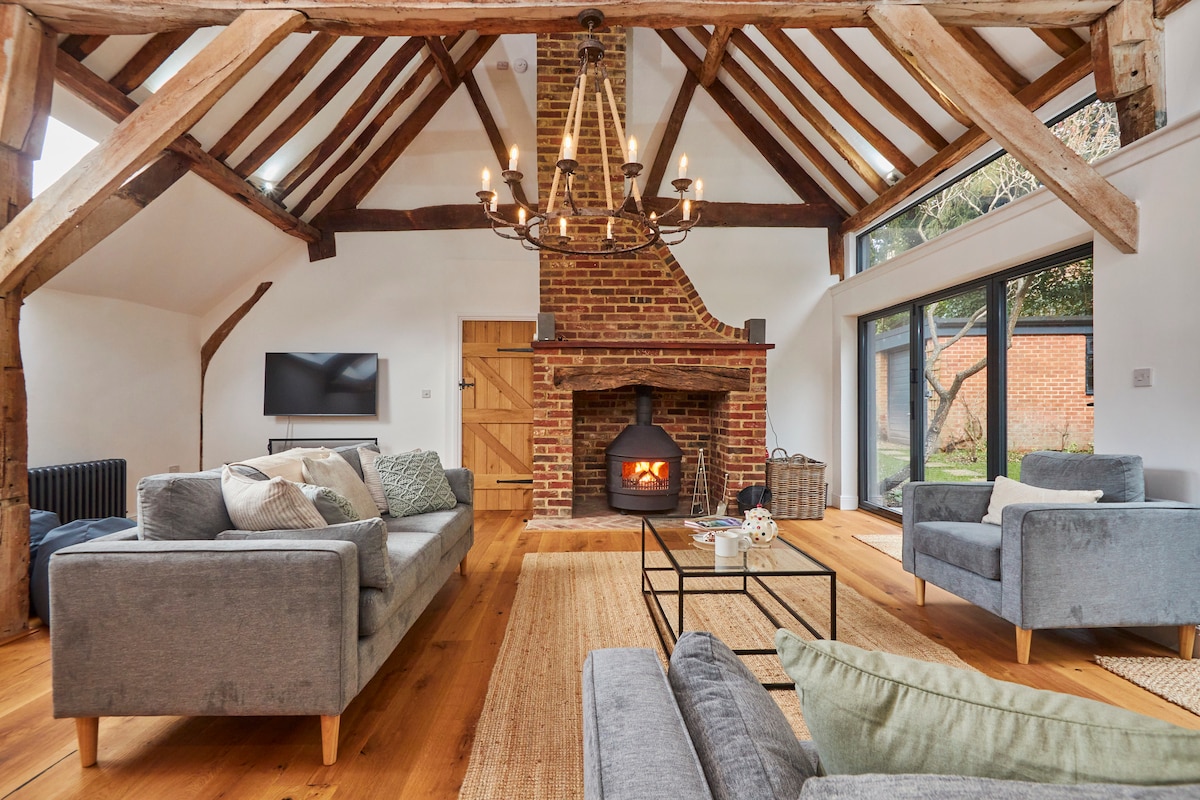 The spacious sitting room features a vaulted ceiling with original beams, enhancing its charm. A cozy fireplace is centrally located, surrounded by comfortable seating. Large windows let in natural light and open to the garden, while a flat-screen TV is positioned on the wall.