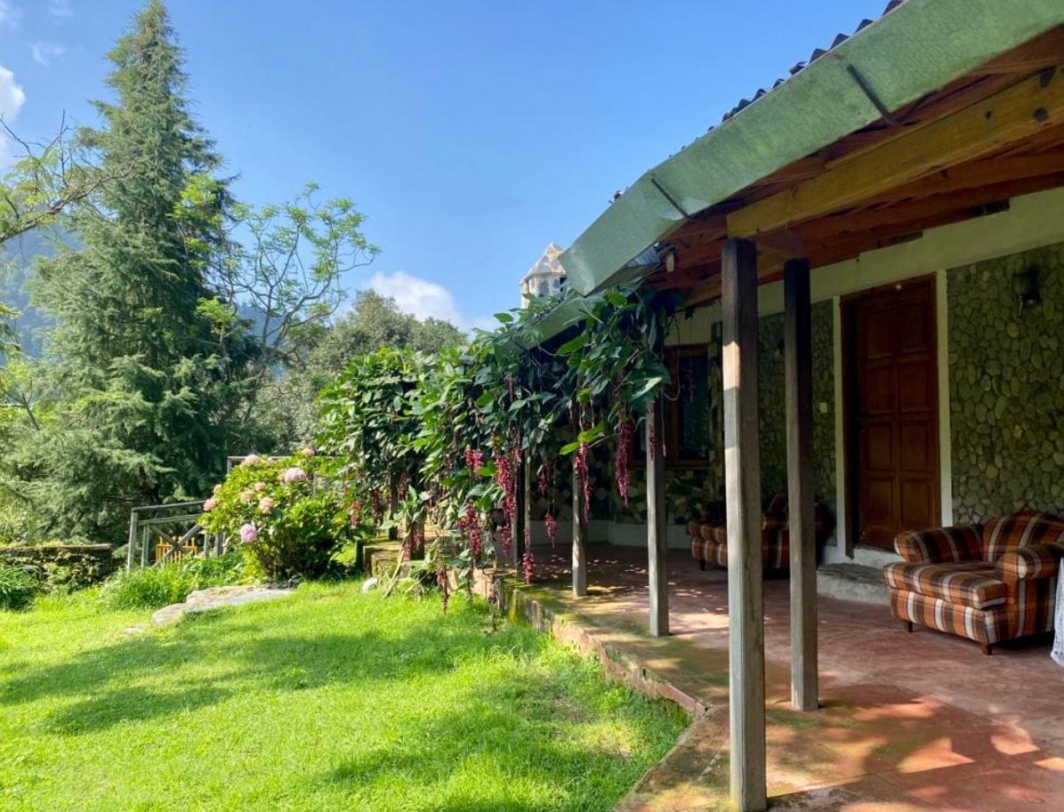 A spacious outdoor area features a green lawn bordered by colorful floral plants and shrubs. The cottage is partially visible, showcasing wooden doors and a stone exterior. Comfortable seating arrangements include patterned chairs under the shade of a roof supported by wooden beams.