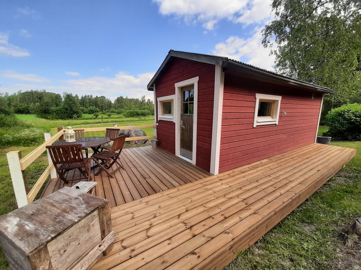 A rustic cabin with red wooden siding is situated on a spacious wooden deck. The deck features a dining table with six wooden chairs, surrounded by greenery. Large windows are visible on the cabin, offering natural light to the interior.