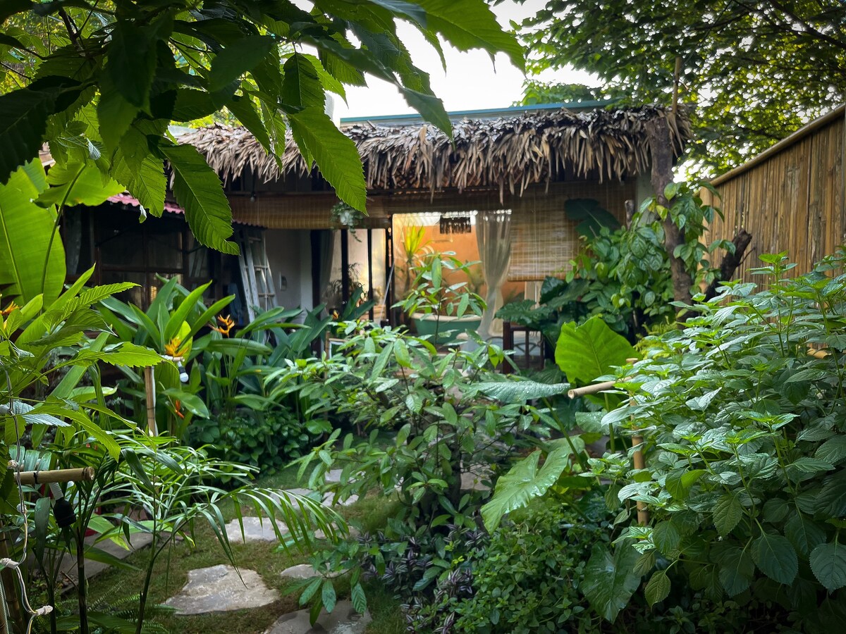 A lush green garden is framed by tropical plants and foliage, leading towards a thatched-roof structure. Natural pathways are visible among the dense greenery, enhancing a sense of seclusion. The entrance features warm light, inviting exploration of the hidden retreat.