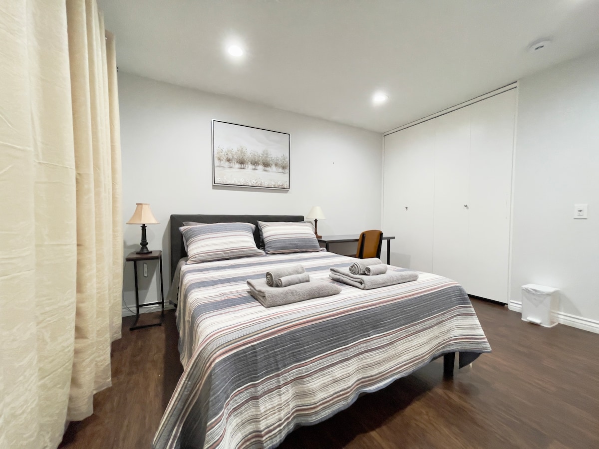 A private room is presented featuring a spacious bed with a striped blanket. Two neatly arranged towels are placed at the foot of the bed. A desk with a chair is positioned against the wall, accompanied by soft lighting from a bedside lamp. A large closet is visible in the background.