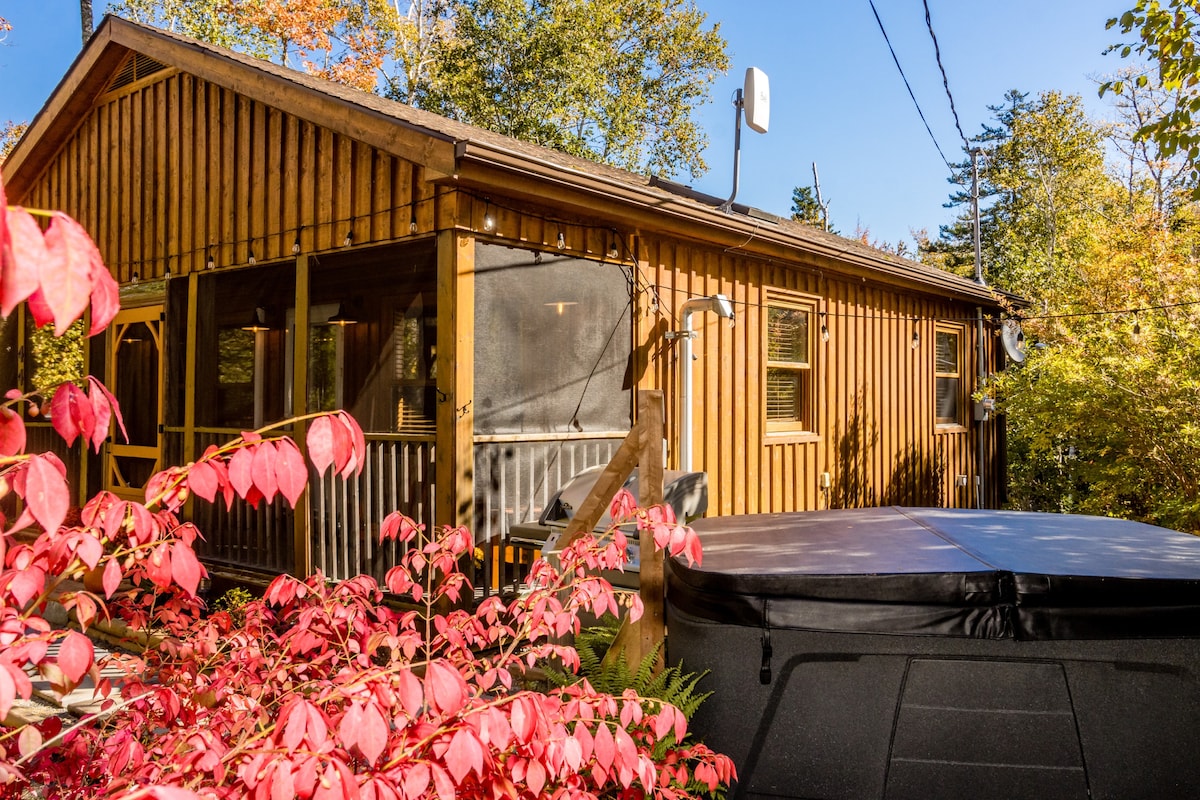 The exterior of the cabin is adorned with natural wood siding, complemented by a screened-in porch. A hot tub is visible on the deck, surrounded by vibrant foliage showcasing autumn colors, creating a serene and secluded atmosphere.
