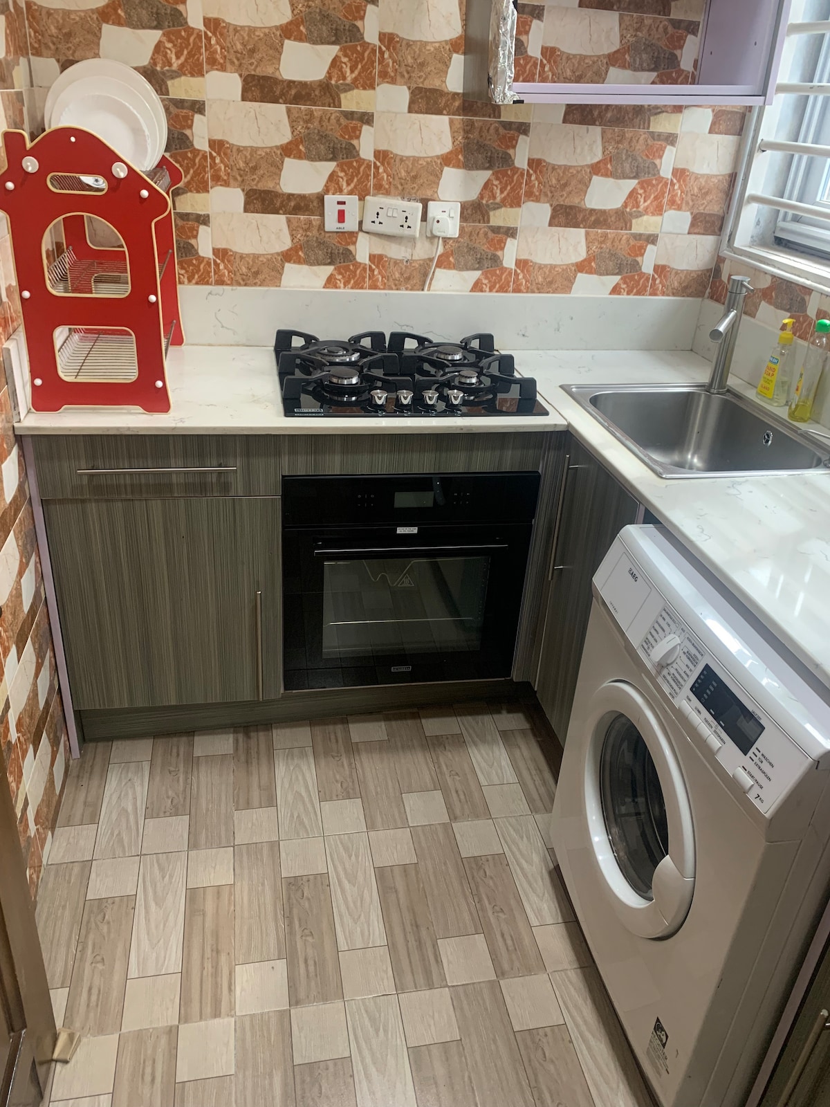 A well-equipped kitchen features modern appliances, including a gas stove and an oven. A washing machine is installed, and a double sink is positioned nearby. The cabinetry is a sleek gray, complemented by a stylish countertop and a patterned backsplash.