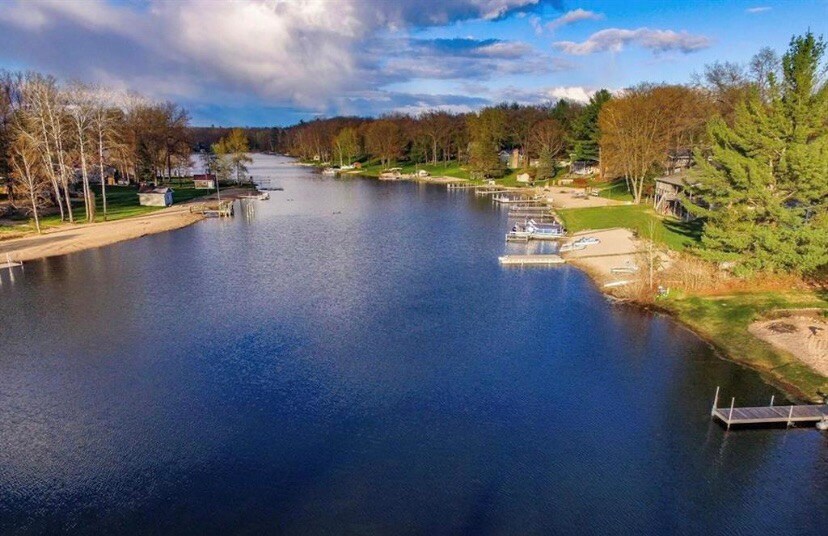 A serene lake view reveals calm waters reflecting the sky, bordered by tree-lined shores. Several docks and boats are anchored along the shoreline, with sandy areas visible, creating a peaceful atmosphere for outdoor activities.