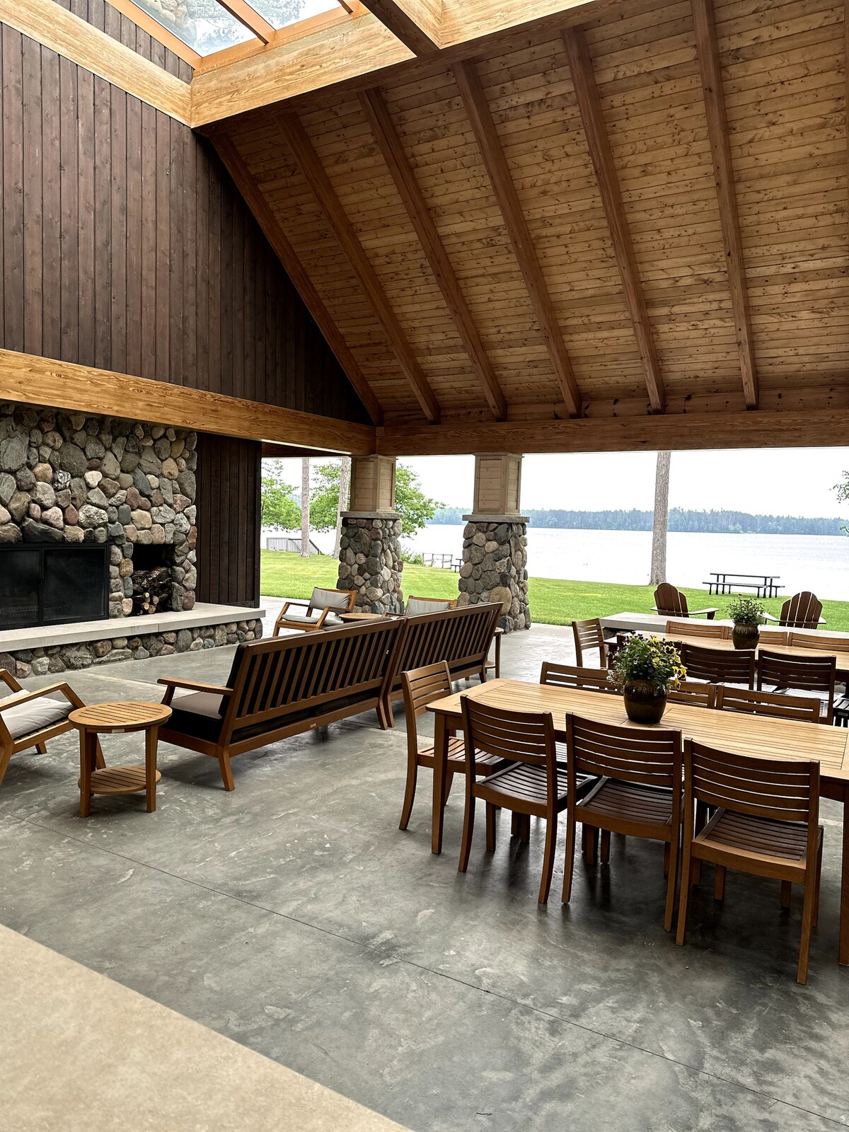 An open-air living space features a high wooden ceiling and a stone fireplace. Comfortable wooden seating arrangements, including a long dining table, provide ample space for gatherings. Large windows offer a view of the lake and surrounding greenery, enhancing the inviting atmosphere.