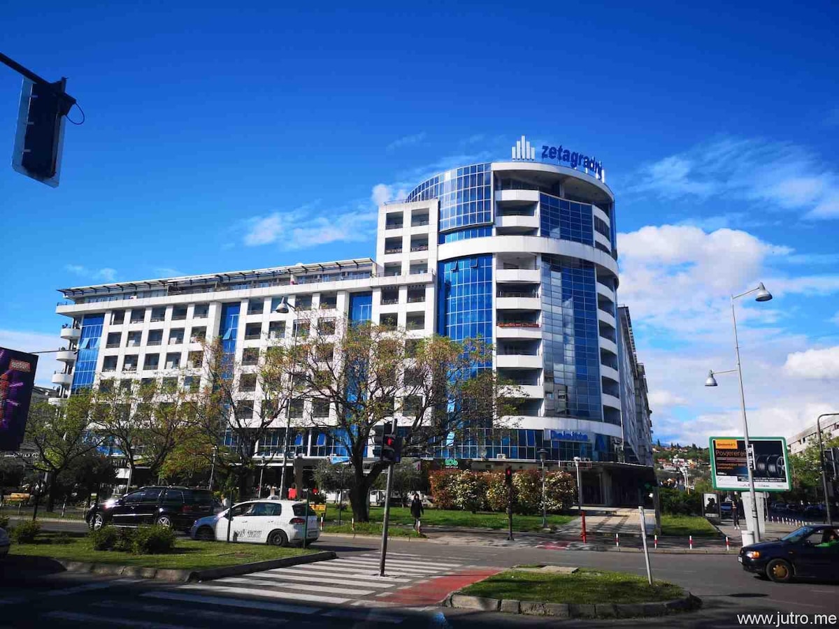 A modern building features a sleek design with a combination of glass and concrete. Large windows reflect the sky, while trees line the surrounding area. The location is busy, with traffic signals and pathways visible, indicating an urban environment.