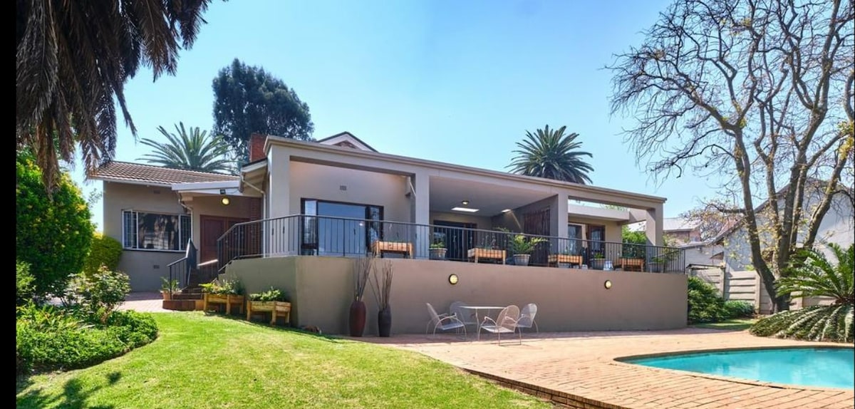 A spacious villa is set on a lush green lawn, featuring a large patio with outdoor seating. The inviting pool glistens under the sunlight, surrounded by well-maintained gardens. Palm trees and flowering plants provide a serene atmosphere, enhancing the outdoor living space.