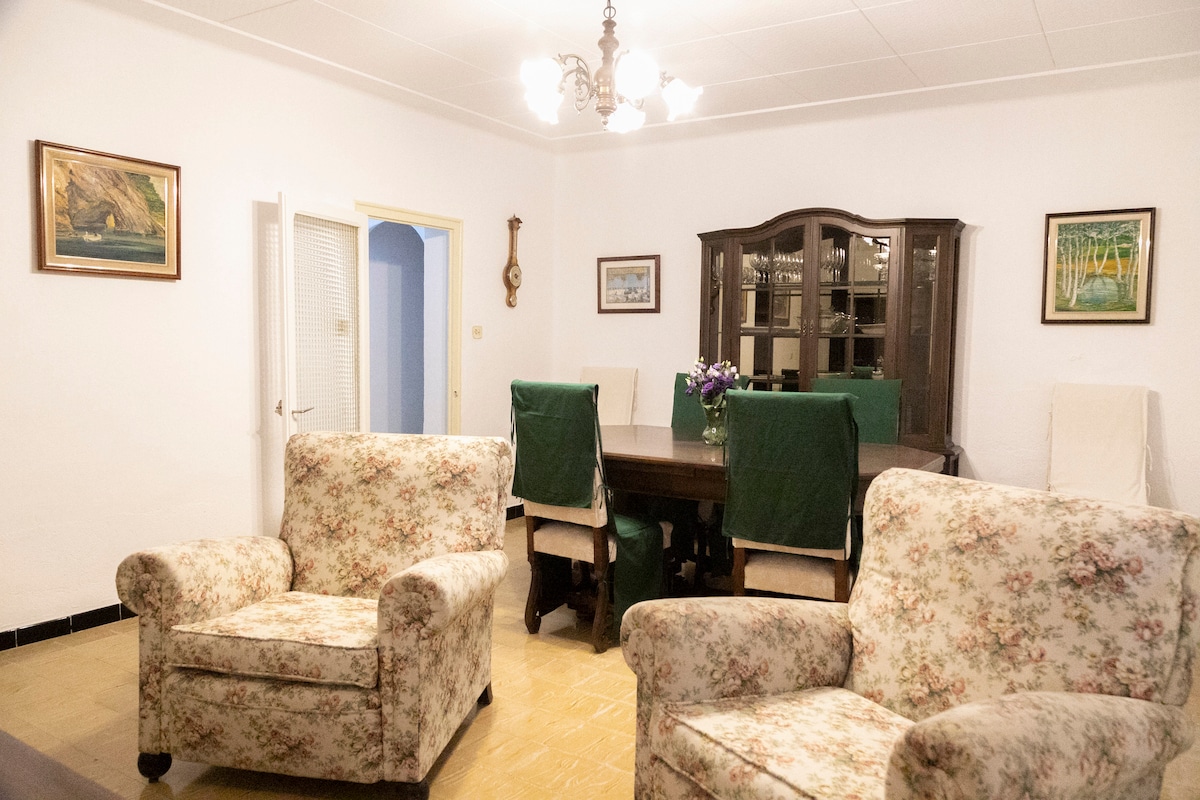 A spacious living area is furnished with floral-patterned armchairs. A wooden dining table, surrounded by green-covered chairs, is centrally positioned. Soft lighting from the chandelier illuminates the room, enhancing the warmth of the light-colored walls adorned with framed artwork.