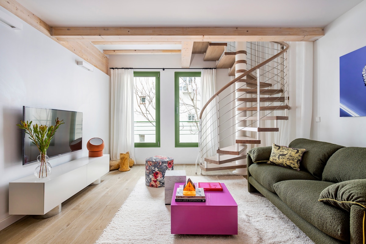 A stylish living area features a green sofa and a vibrant purple coffee table. Natural light filters through large green-framed windows, illuminating the space, which is complemented by a spiral staircase leading to an upper level. A modern television is mounted on a minimalist media unit.