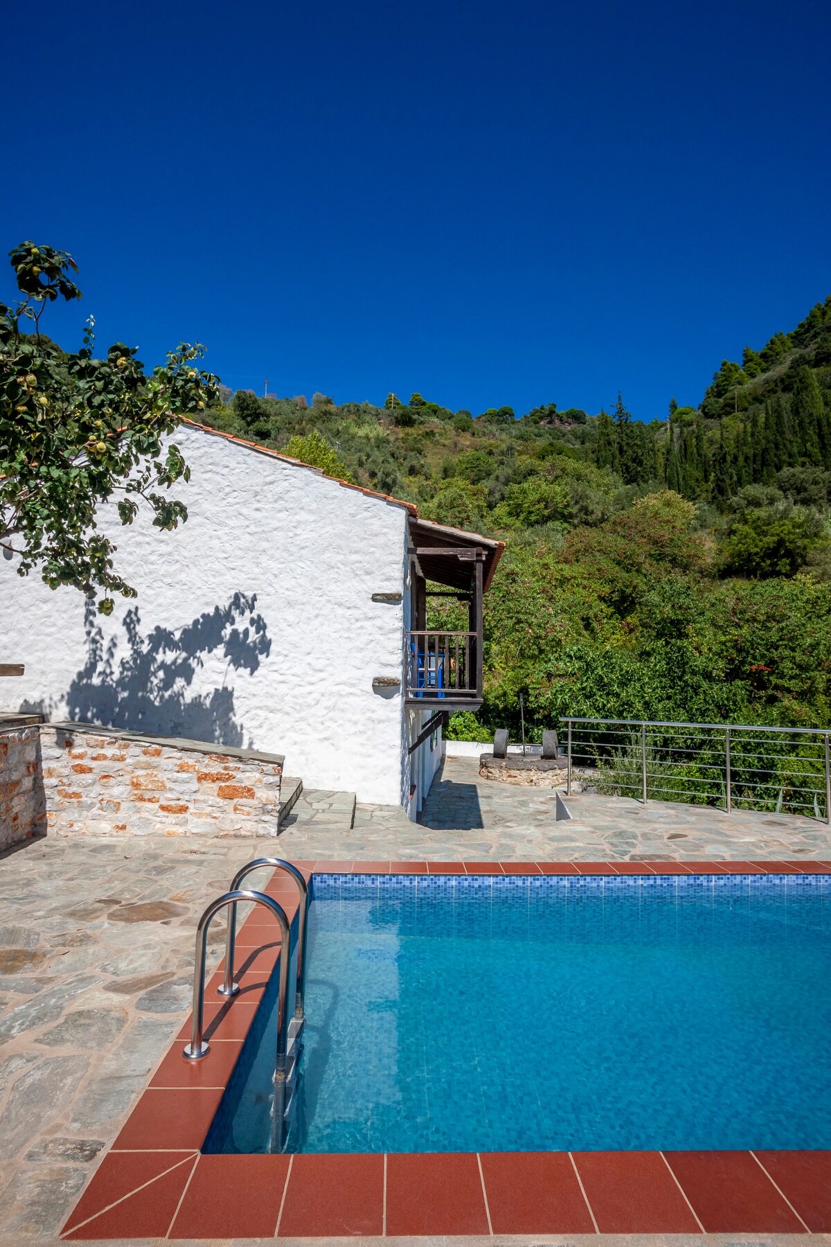 A refreshing swimming pool is framed by a terracotta border, with sparkling blue water reflecting the clear sky. The adjacent white building features a wooden balcony overlooking lush greenery. Stone pathways lead around the pool area, enhancing the natural landscape.