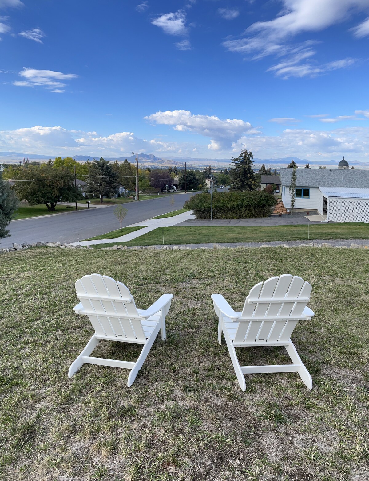 Two Adirondack chairs are positioned on a grassy area, overlooking a scenic view of the neighborhood and distant hills. The sky is partly cloudy, with blue and white tones enhancing the overall tranquil setting.