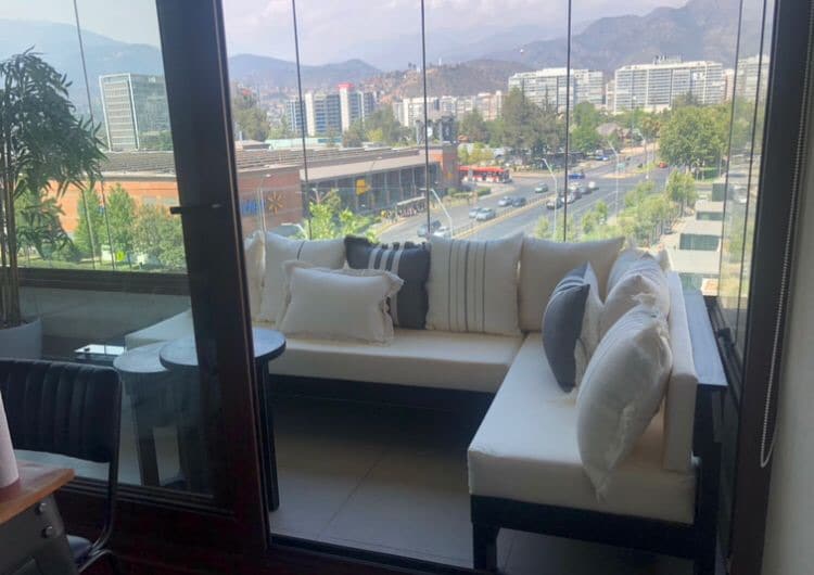 A spacious seating area features a modern outdoor couch adorned with decorative pillows. Large glass walls provide panoramic views of the surrounding cityscape and mountains, allowing plenty of natural light to brighten the area.