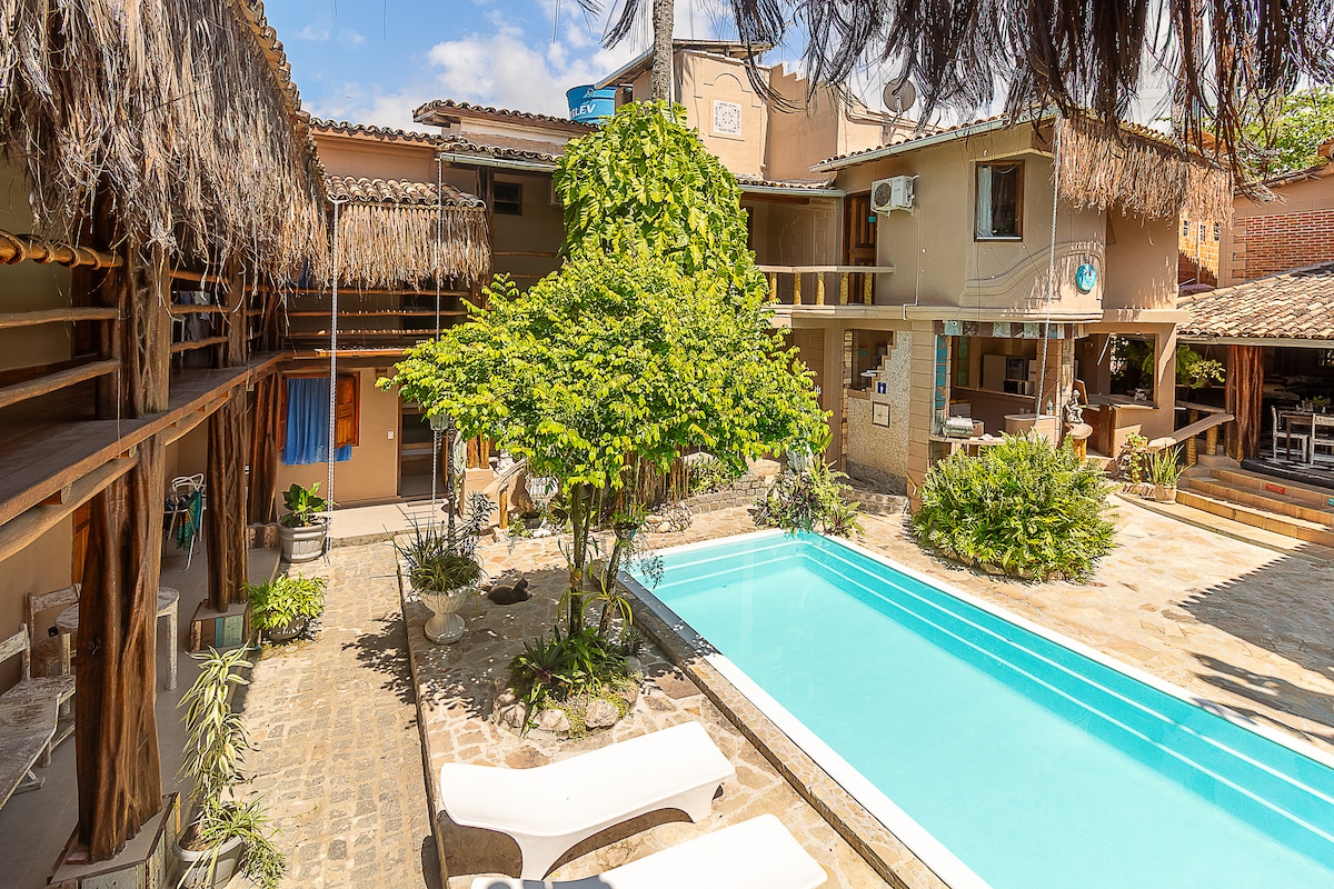 A central pool surrounded by a stone patio is enhanced by lush greenery and a tall tree. Sun loungers are positioned adjacent to the pool, and a common area is visible with thatched roofs and various plants, creating a relaxed outdoor environment.